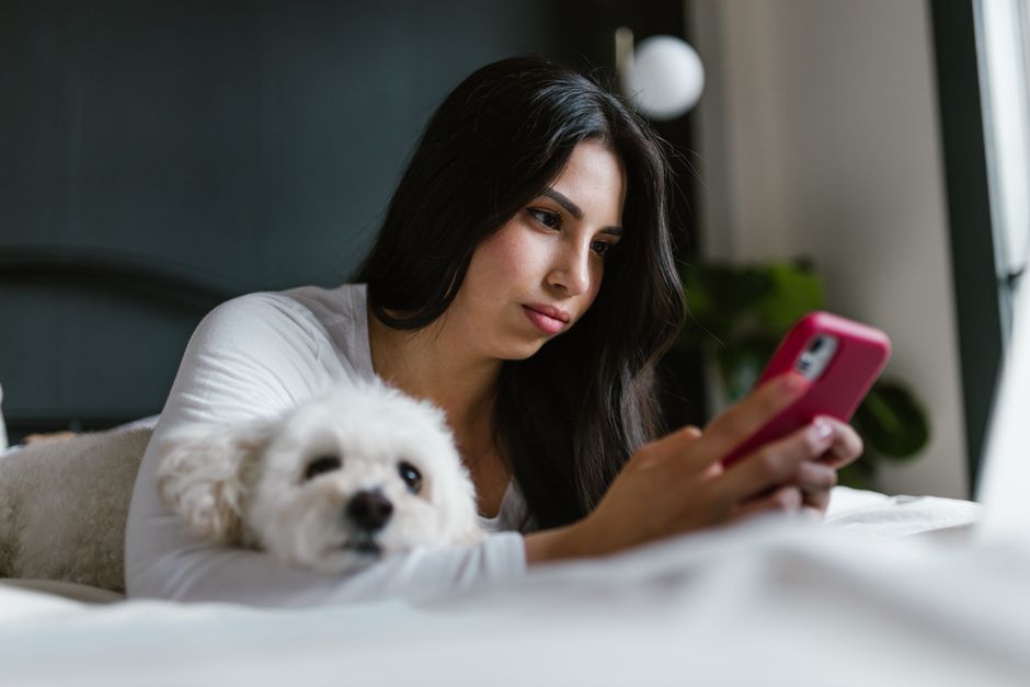 Woman lying in bed using a smartphone with her pet dog beside her